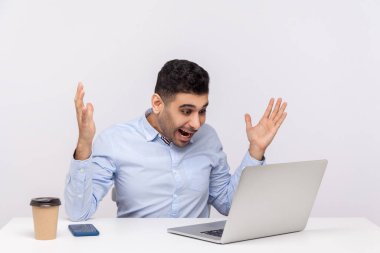 Vay canına, inanılmaz! Şaşırtıcı derecede mutlu bir işadamı ofis yerinde oturmuş, şaşkınlıkla el kaldırıyor, şok olmuş bir ifadeyle dizüstü bilgisayarına bakıyor, video konuşmasında konuşuyor. Stüdyoda çekim izole edildi