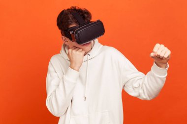 Rastgele giyinmiş ciddi konsantre bir adam beyaz kapüşonlu ve sanal gerçeklik kulaklıklı yumrukları boksa hazır, online oyun oynamaya hazır. Turuncu arka planda kapalı stüdyo çekimleri