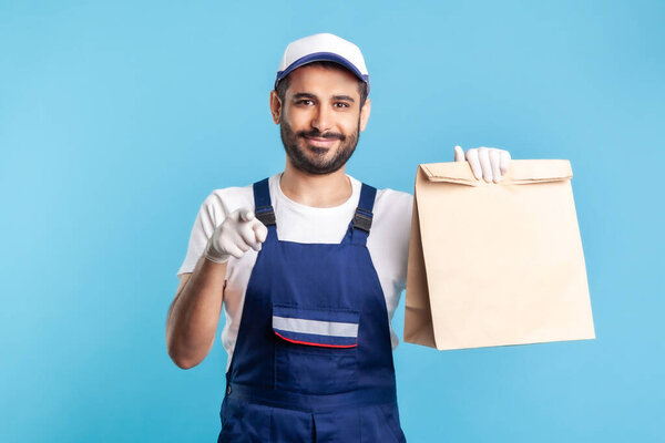 Hey you! Portrait of cheerful bearded handyman in overalls and gloves carrying parcel, pointing to camera. Courier delivering food in paper bag, post mail services. indoor studio shot, isolated