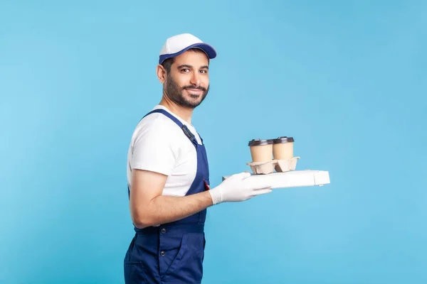 Delivery service. Side view, happy courier in overalls holding coffee and pizza box, wearing safety gloves offering drinks food and smiling to camera. indoor studio shot isolated on blue background