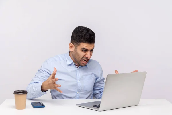 Irritated businessman sitting office workplace, looking at laptop with furious expression, shouting angry while having online conversation, video call. studio shot isolated on white background