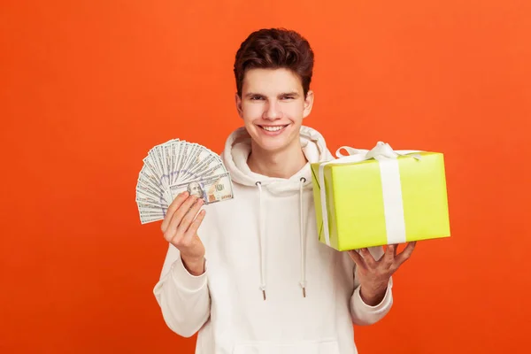 笑顔カジュアルなパーカーのギフトボックスとお金の束を保持し キャッシュバック 銀行融資で休日のショッピングで満足した若い男 オレンジの背景に隔離された屋内スタジオショット — ストック写真