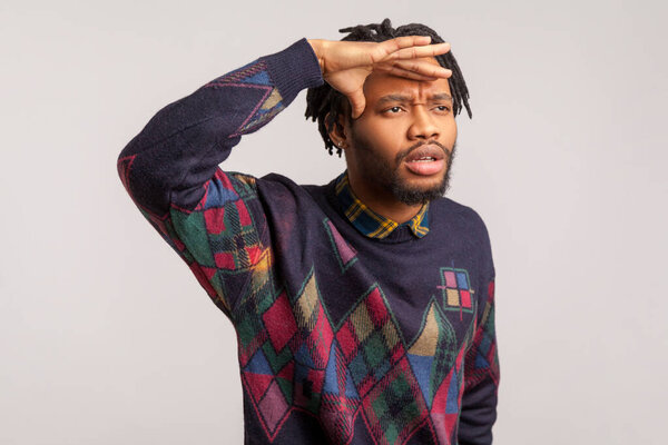 Concentrated african man with dreadlocks holding hand over head looking far away, trying to see future and perspectives. Indoor studio shot isolated on gray background