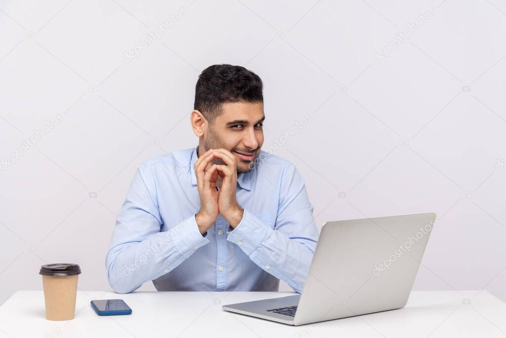 Sneaky scheming businessman sitting office workplace with laptop on desk, clasping hands and smirking mysteriously, having tricky cunning plan in mind. indoor studio shot isolated on white background