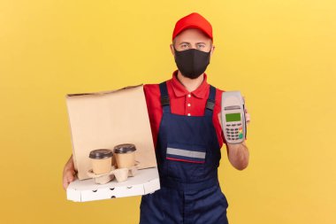 Mavi üniformalı ve hijyenik koruyucu maskeli bir kurye postu, pizza kutulu kahve ve karantinada teslim sırasında güvenlik ödemesi tutuyor. Sarı arka planda kapalı stüdyo çekimleri