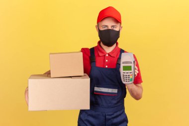 Teslimatçı, yüzünde koruyucu maskeyle karton kutular ve kontak olmayan ödemeler için postaneyi gösteriyor, karantinaya teslim ediliyor. Sarı arka planda kapalı stüdyo çekimleri