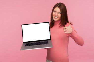 Kahverengi saçlı, pembe kazaklı, dizüstü bilgisayarlı, boş ekranlı, mutlu ve online uygulamadan memnun bir kadın. Pembe arka planda kapalı stüdyo çekimleri