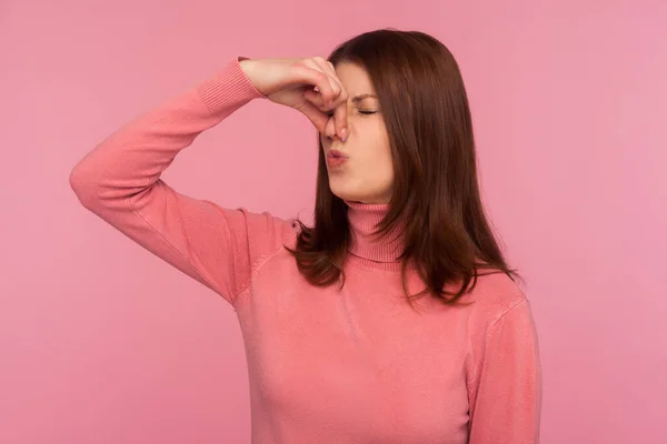 Unhappy confused brunette woman grabbing nose with fingers feeling unpleasant smell, shocked with disgusted fart. Indoor studio shot isolated on pink background