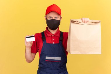 Mavi üniformalı, kırmızı tişörtlü ve şapkalı hijyenik koruyucu maskeli, elinde kağıt çanta ve kredi kartı olan teslimatçı. Nakitsiz ödemeler. Sarı arka planda kapalı stüdyo çekimleri
