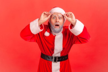Son derece şaşırmış Noel Baba 'nın portresi kameraya bakarken büyük merak uyandıran gözlerle alnına gözlük takarken, bayramlar yaklaşırken. Kırmızı arkaplanda kapalı stüdyo çekimleri
