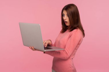 Pembe kazaklı sürpriz esmer kadın dizüstü bilgisayara büyük gözlerle bakıyor, iş gereksinimleri karşısında şokta, son teslim tarihi. Pembe arka planda kapalı stüdyo çekimleri