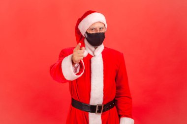 Tıbbi koruyucu maskeli ciddi bir noel baba kamerayı işaret ediyor, salgın hakkında uyarıyor, covid-19 önleme. Kırmızı arkaplanda kapalı stüdyo çekimleri
