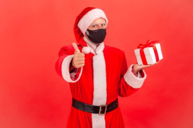 Koruyucu maskeli, yüzünde kırmızı fiyonklu hediye kutusu olan ve bayram hediyelerini seven tatmin olmuş Noel Baba. Kırmızı arkaplanda kapalı stüdyo çekimleri