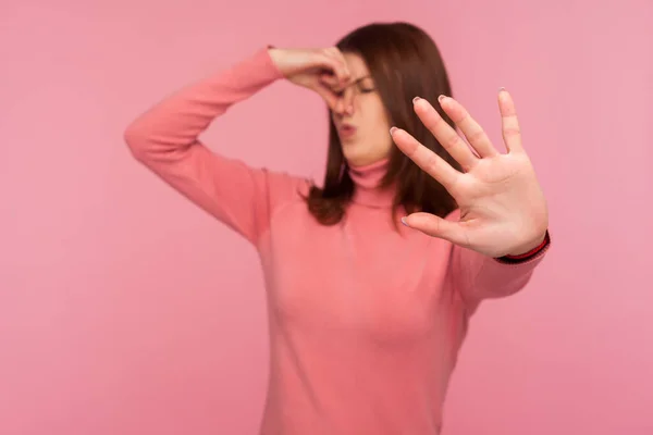 Confused brunette woman holding breath with fingers on nose showing stop gesture, dont come closer, shocked with disgusting stink. Indoor studio shot isolated on pink background