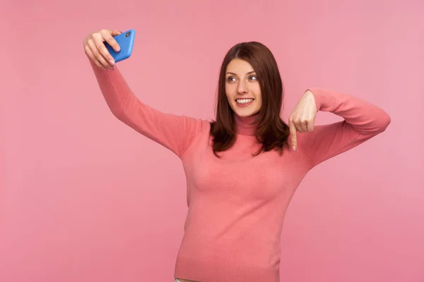 Young woman blogger in pink sweater recording video message to her admirers on smartphone asking to subscribe and rate her posts in social network. Indoor studio shot isolated on pink background