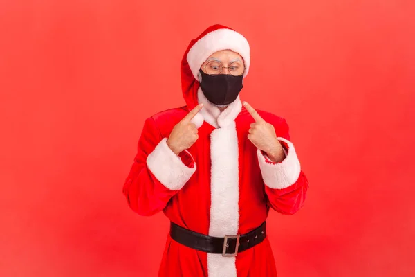 Scared funny man in santa claus costume pointing fingers and trying to look at protective hygienic mask on his face, tired of covid-19 epidemic. Indoor studio shot isolated on red background