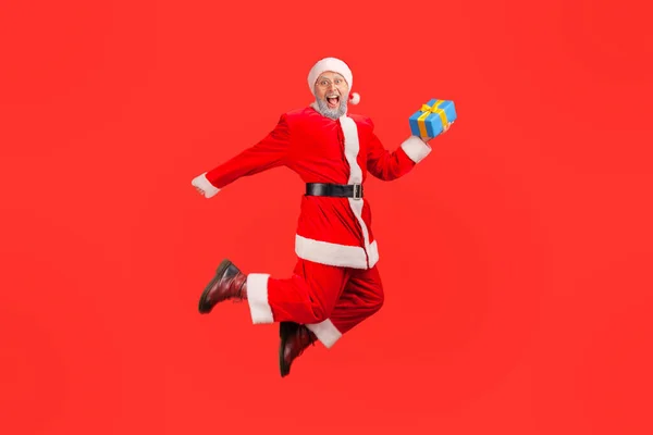 非常快乐的圣诞老人拿着礼品盒跳得高高的 面带微笑地看着相机 对寒假感到满意 在红色背景下隔离的室内拍摄 — 图库照片