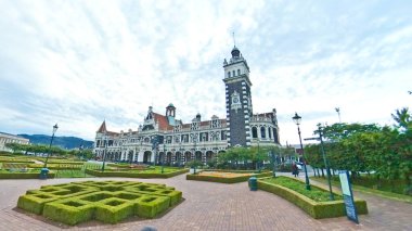 Uzun saat kulesi olan çarpıcı bir tren istasyonu resmi bir bahçenin ve tuğla bir meydanın arkasında duruyor. 