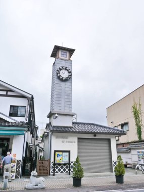 Güvenlik kamerası olan uzun ve modern bir saat kulesi aşağıdan görülüyor. Binada Japonca karakterler var. 