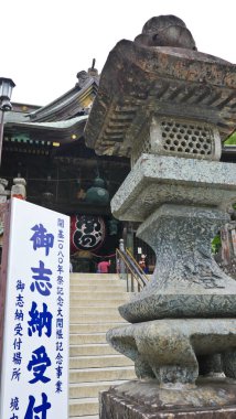 Büyük, yıpranmış bronz bir fener merkezde duruyor, daha küçük, süslü taş fenerlerle çevrili, arka planda yemyeşil ağaçlar var. 