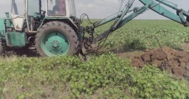 Bir kazıcı, sahadan çıkabilecek yangını önlemek için hendek kazıyor. Traktör sürücüsü sahada çalışıyor. Çukur kazmak
