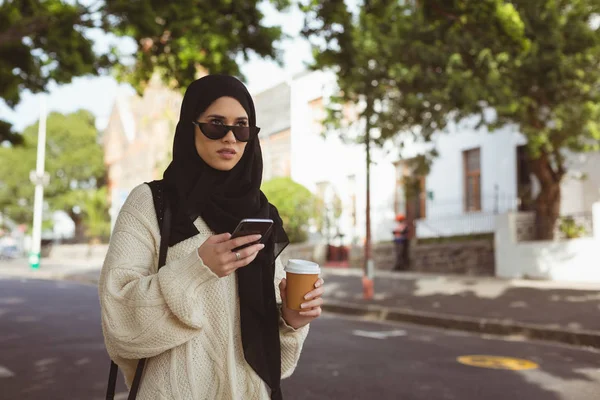 ヒジャーブ女性ストリートでコーヒー カップを保持している間携帯電話を使用して — ストック写真