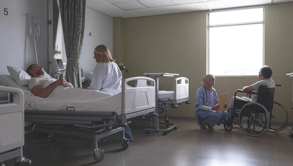 病院の病棟でベッドに横たわっている白人患者と相互作用する白人女性医師の側面図 ヒジャーブの混合人種女性看護師が 窓の横に車椅子で先輩の混合人種男性患者と医療フォルダーを議論する — ストック写真