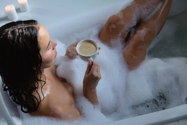 若い白人ブルネットの女性がコーヒーを飲みながら足を上げて泡風呂に座っている高い眺め — ストック写真