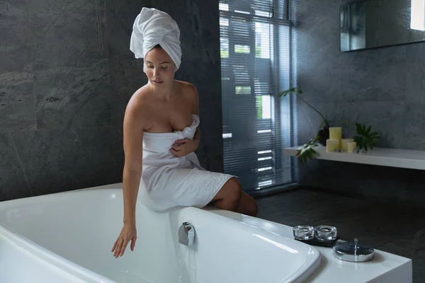 一个年轻的白种妇女穿着浴巾 头发裹着毛巾 坐在浴缸边 在现代浴室里摸着水的正面景色 — 图库照片