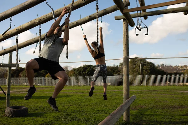在训练营训练课上 一名年轻的白种人男子在户外健身房的攀爬架上用绳子吊起 背景中一名妇女从绳子上吊起 — 图库照片