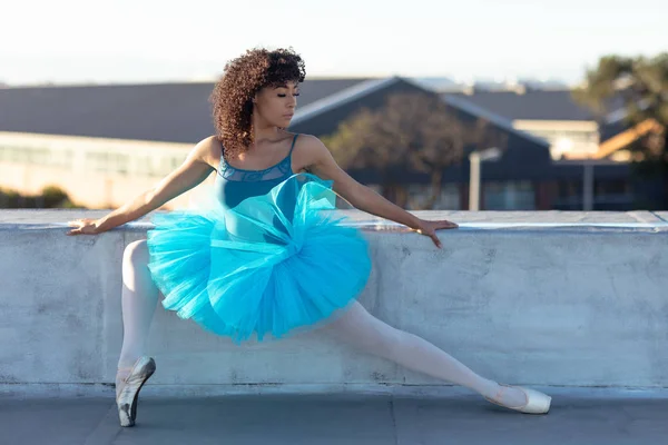 バレエの位置を持ち 都会の建物の屋上を見下ろす青いチュチュを着た若い混合人種の女性バレエダンサーの正面図 — ストック写真