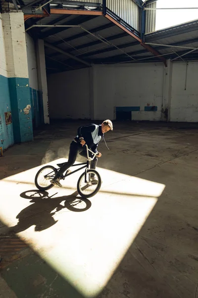 放棄された倉庫でトリックを練習しながら Bmxバイクの日光のシャフトで停止するために横にスライドする若い白人男性のサイドビュークローズアップ — ストック写真