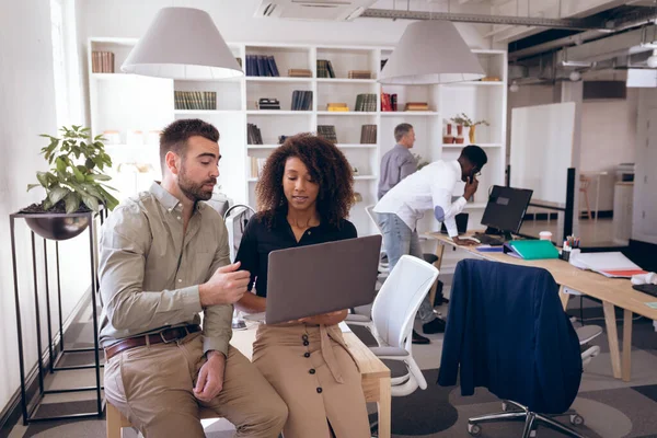 Eine Geschäftsfrau mit gemischter Rasse und ein kaukasischer Geschäftsmann arbeiten in einem modernen Büro, benutzen einen Laptop und unterhalten sich, während ihre Geschäftskollegen im Hintergrund arbeiten. — Stockfoto