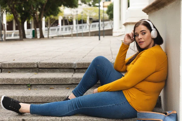 Curvy Caucasian woman out and about in the city streets during the day, sitting on steps, leaning back and listening to music on headphones, with historical building in the background — Stock Photo