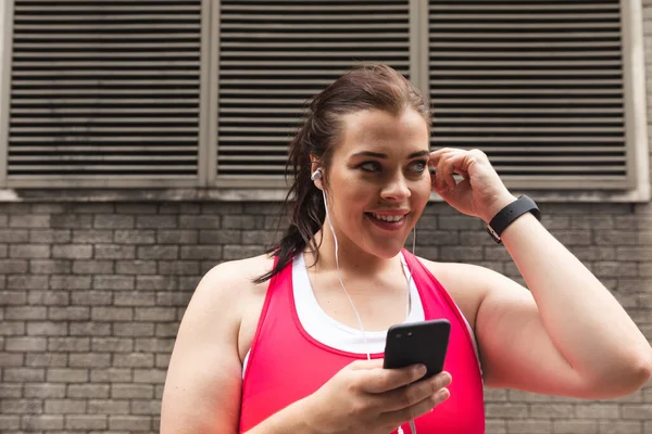 Sonriendo curvilínea mujer caucásica con el pelo largo y oscuro usando ropa deportiva haciendo ejercicio en una ciudad, usando su teléfono inteligente con auriculares, una pared de ladrillo en el fondo - foto de stock