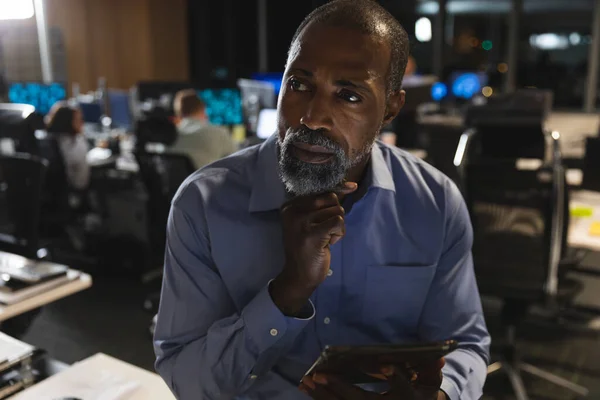 Afroamerikanischer Geschäftsmann arbeitet spät abends in einem modernen Büro, sitzt auf einem Schreibtisch, benutzt einen Tablet-Computer, hält sein Kinn und denkt nach.. — Stockfoto