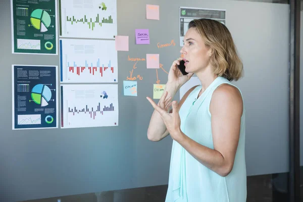 Smart casually dressed Caucasian businesswoman talking on smartphone, a board with charts and information on it in the background. Creative business professional working in a busy modern office. — Stock Photo