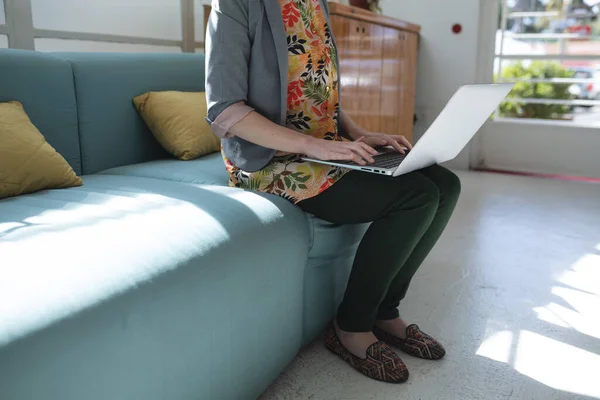 Caucasian female business creative sitting on sofa in an office using her laptop. Health and hygiene in workplace during Coronavirus Covid 19 pandemic. — Stock Photo