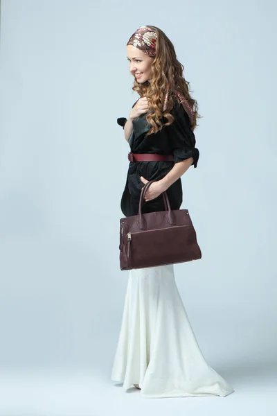 Stylish beautiful woman wearing a scarf on her head, holding a fashionable handbag in her hand. Studio photography on a blue background. Vertical orientation