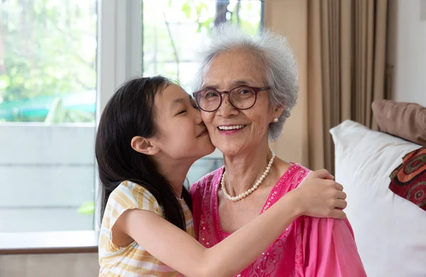 Portrait of cute little girl and her beautiful grandma sitting on sofa at home ,Asian girl kissing her smiling granny in cheek