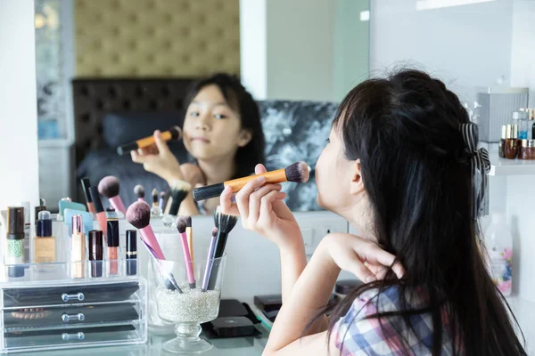 Happy asian little girl, adding some color to my cheeks and applying foundation powder or blush with makeup brush,side view of cheerful little girl applying make-up and looking at her reflection in mirror while sitting at the dressing table at home