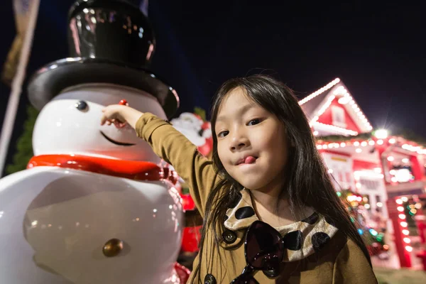 Asijská Holčička a sněhulák stojící v poli, noční pohled, v pozadí domu a vánoční strom v novém let šťastné dítě v Prázdninové aktivity — Stock fotografie