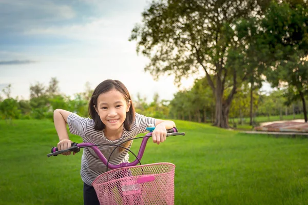 Asiatisk tjej tittar på kameran, leende med en söt på cykel i parken utomhus, barn motion i naturen på morgonen, hälsosam livsstilskoncept — Stockfoto