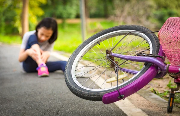 Азиатская девочка, сидящая на дороге с легкой болью в ноге из-за падения велосипедной аварии, велосипед падает перед ребенком в парке, падающий велосипед, концепция ДТП — стоковое фото
