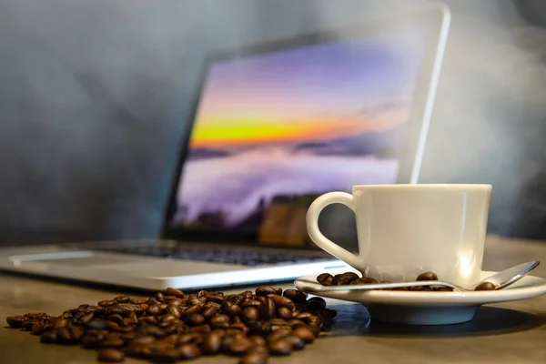 Café tostado y café expreso en una taza de café caliente con vapor y humo caliente, fondo negro, lugar de trabajo en una cafetería, una taza de café al lado de una computadora portátil, planeando un viaje — Foto de Stock