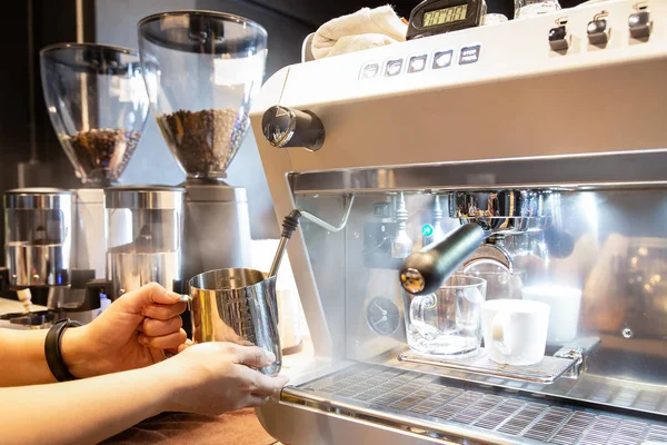 Woman hands holding stainless steel milk pitcher,girl barista steaming milk in the stainless steel milk pitcher for hot cappuccino with coffee machine at the coffee shop