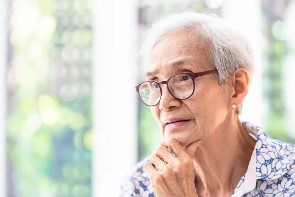 Asian elderly woman in glasses,thinking with hand on chin in her home,senior woman smiling feeling happy