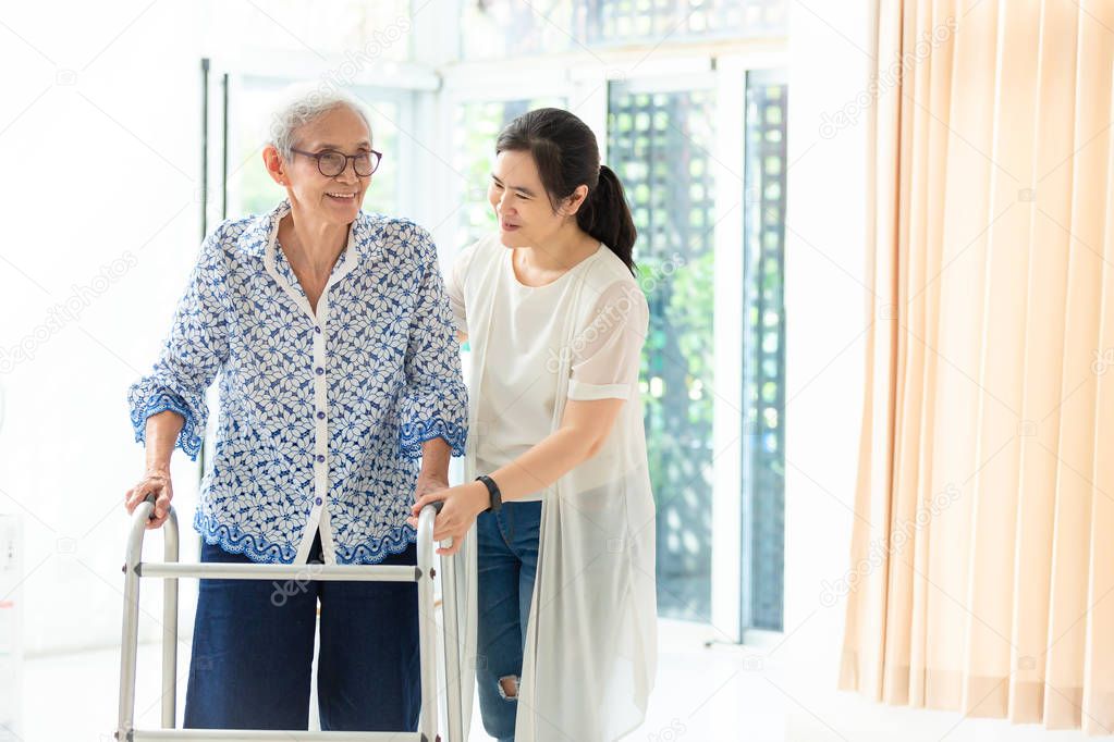 Asian young woman helping senior woman in using walker during  rehabilitation, close up of carer supporting her elderly grandmother with walking and exercising at home