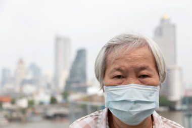 Yüz maskesi koruması olan Asyalı yaşlı kadın, arka plan olarak şehir binasında hava kirliliği nedeniyle yüz maskesi takan yaşlı kadın,Tıbbi maskeile hasta yaşlı insanlar;kirlilik kavramı, toz alerjisi ve sağlık