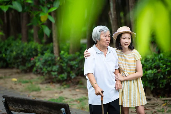 Niña asiática apoyando a la mujer mayor con bastón, feliz abuela sonriente y nieta en el parque, ancianos caminando ejercicio para la salud, concepto de familia; verano y vacaciones — Foto de Stock